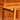 Close-up of a wooden chair with armrests and smooth edges, finished naturally. In the softly blurred background is a Mid-Century Noblett Drinks Trolley – Removable Tray & Original Castors (Revive), echoing the chair’s elegant style.
