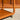 Close-up of the Mid-Century Noblett Drinks Trolley – Removable Tray & Original Castors (Revive), showing its rounded edges and castor, sunlit wood grain on a concrete floor. A blurred wooden cabinet appears in the background.